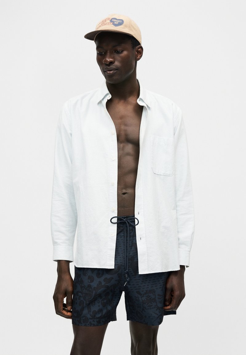 Junger Mann mit hellbeiger Kappe, aufgeknöpftem weißen Hemd mit langen Ärmeln und dunkel gemusterten Shorts vor einem schlichten hellen Hintergrund.