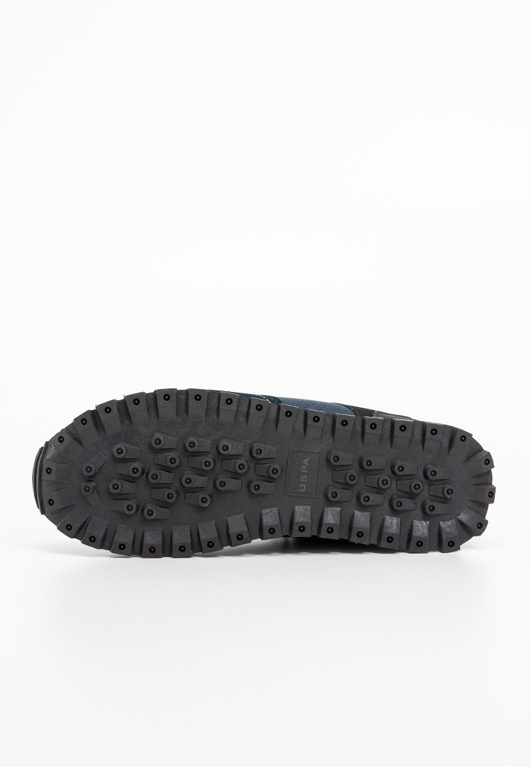 Suela de zapato de goma negra con profundas ranuras para tracción. Nota el texto "USPA" y los tacos elevados para agarre. La forma es curvada, adecuada para uso atlético.