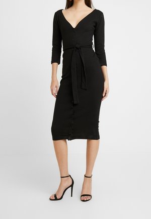 Femme portant une robe midi noire ajustée avec des manches trois-quarts, des boutons devant, une ceinture nouée à la taille et des sandales noires à talons hauts.