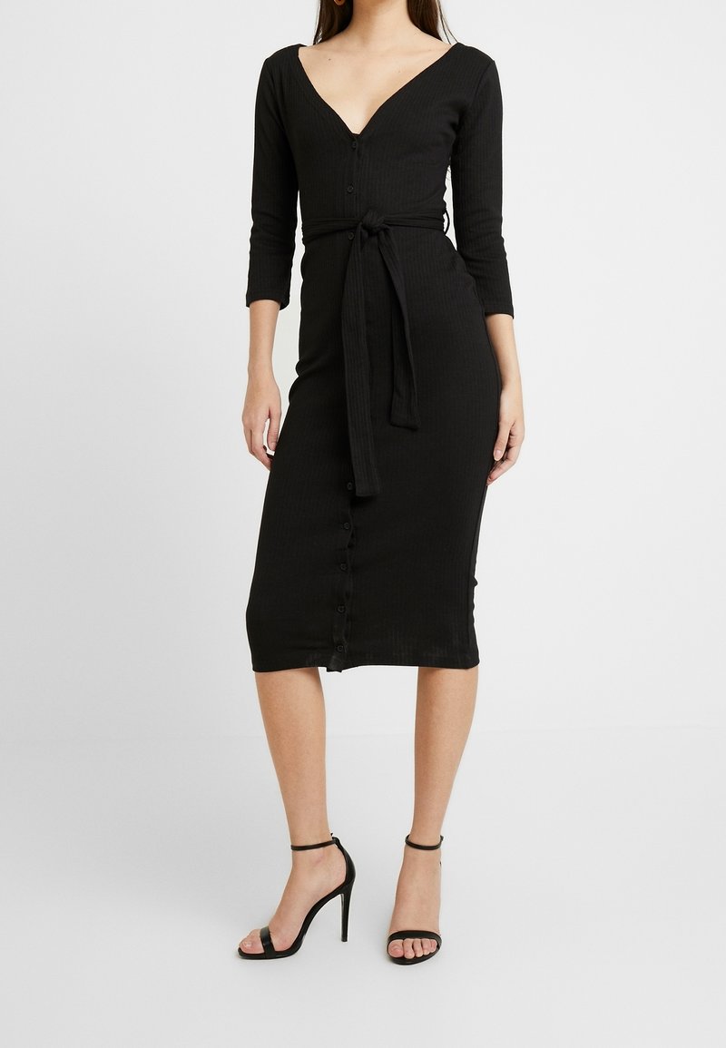 Femme portant une robe midi noire ajustée avec des manches trois-quarts, des boutons devant, une ceinture nouée à la taille et des sandales noires à talons hauts.