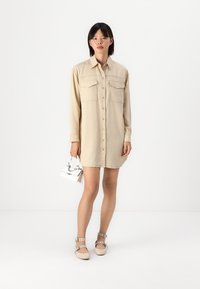 Vestito camicia beige con bottoni, maniche lunghe, due tasche sul petto e una silhouette rilassata, abbinato a scarpe flat pastello e una piccola borsa.