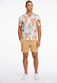 Camisa de manga corta con estampado de hojas tropicales multicolores sobre un fondo blanco. Combinada con pantalones cortos de pana en color beige y zapatillas blancas.
