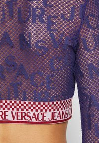Crop top in mesh viola con un motivo testuale intrecciato e una banda a scacchi contrastante in maroon e rosa con la scritta "VERSACE JEANS".