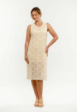 Vrouw met blond haar die een mouwloze beige kanten midijurk en beige sandalen draagt, staat met één hand op haar heup.