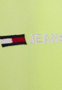 Tessuto giallo neon con la scritta ricamata "JEANS" e un dettaglio logo rettangolare orizzontale rosso, bianco e nero. Texture liscia.