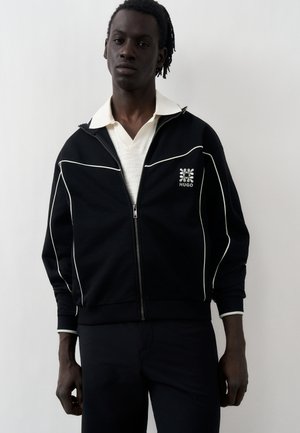 Νέος άνδρας με σκούρο δέρμα και κοντά dreadlocks, φοράει μαύρο μπουφάν με φερμουάρ και λευκές ρίγες και κάτω έχει λευκό πουκάμισο με γιακά.