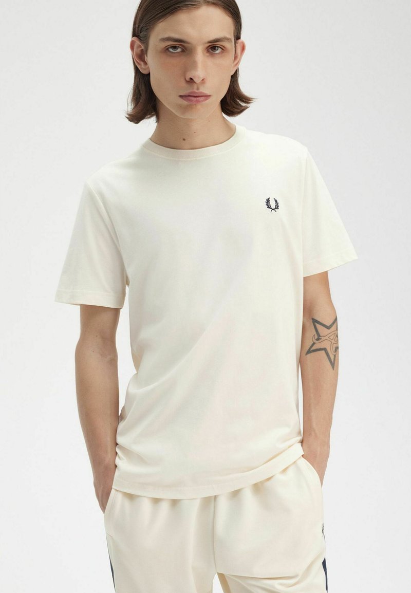 Camiseta de algodón color crema con mangas cortas y escote redondo, que presenta un pequeño logo azul marino en el lado izquierdo del pecho. Diseño sencillo.
