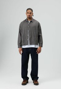 Geruit overhemd in grijs en bruin, gecombineerd met een wit T-shirt eronder en donkerblauwe spijkerbroek. Bruine gestructureerde laarzen maken de look compleet.