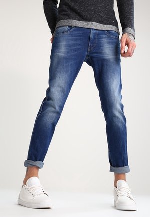 Mand iført op rullede blå jeans, hvide sneakers og en mørk langærmet sweater står med benene fra hinanden på en ensfarvet baggrund.