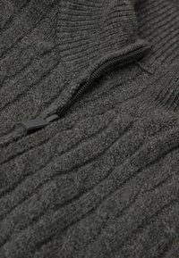 Světle šedý pletený svetr s ribovanou texturou, s předním zipem a hladkým jezdcem. Látka vypadá měkko a teple.