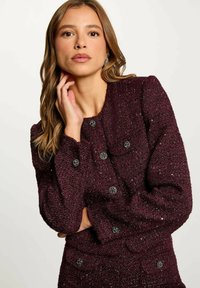 Giacca bordeaux in tessuto con scollatura rotonda, maniche lunghe e bottoni neri decorativi. Presenta due tasche frontali e una silhouette aderente.