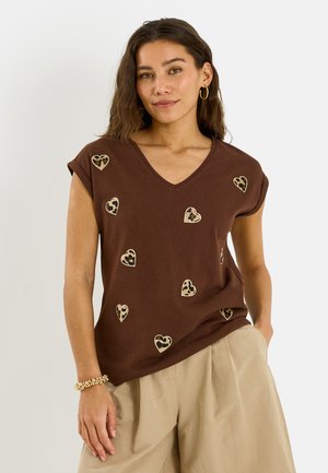 Femme portant un t-shirt marron col en V avec manches retroussées et patchs en forme de cœur imprimés léopard, associé à un pantalon beige à jambes larges.