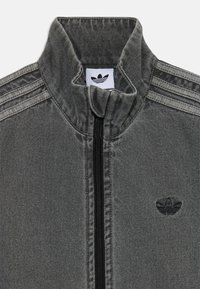 Veste en denim gris avec un col haut, dotée d'une fermeture éclair noire et de trois bandes horizontales sur les épaules. Comprend un logo en patch sur la poitrine.