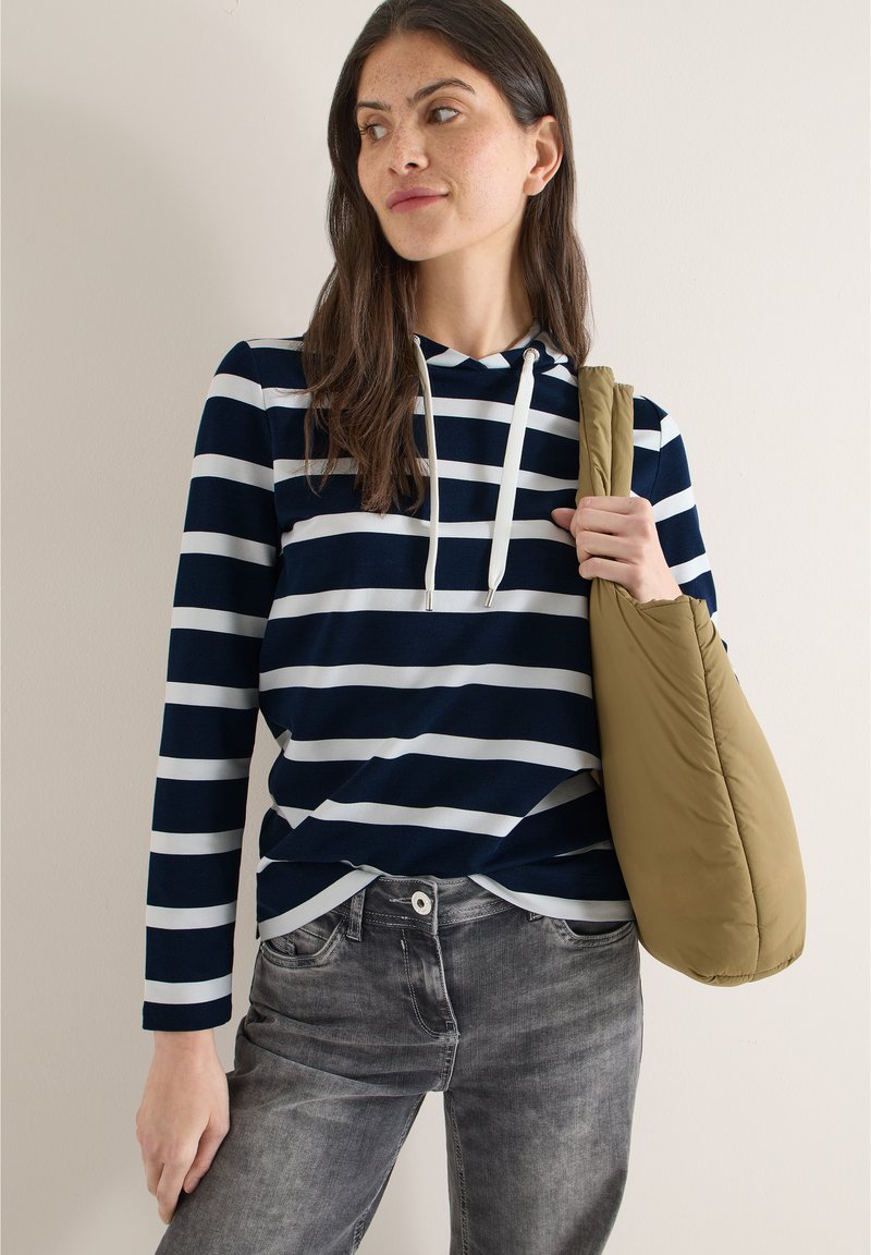 Sweat à capuche à rayures bleu marine et blanc avec manches longues, associé à un jean gris et un sac à bandoulière beige. Texture de tissu lisse et douce.