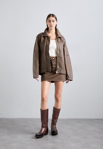 Brun skinnjakke i oversized stil, hvit cropped topp, brun skinn miniskjørt og brune høye støvletter med svarte detaljer. Jevn tekstur.