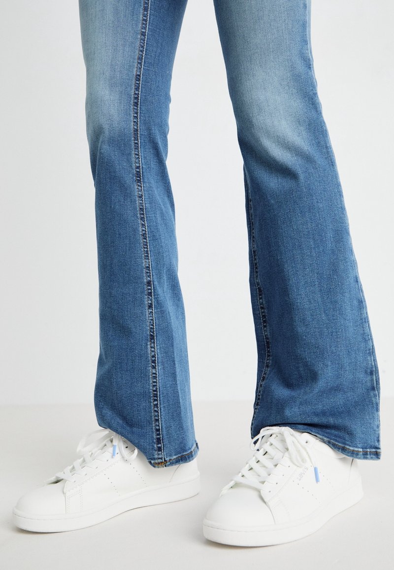 Jambes portant un jean flare bleu et des baskets en cuir blanc à lacets, debout sur un sol et un fond blancs unis.