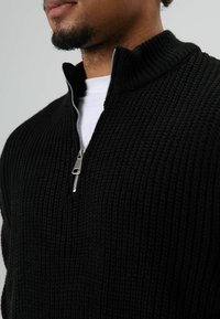 Zwarte gebreide trui met een hoge kraag, zilveren ritssluiting bij de hals, met een gestructureerd patroon. Gedragen over een wit overhemd.