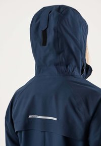 Person iført navyblå vandtæt jakke med hætte trukket op, der viser ryggen med en reflekterende "Swedemount"-logo og en ventilationsklap.