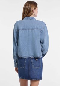 Giubbotto di jeans azzurro chiaro con design cropped, maniche lunghe, polsini con bottoni e un yoke sul retro. Indossato sopra una gonna di jeans blu scuro.