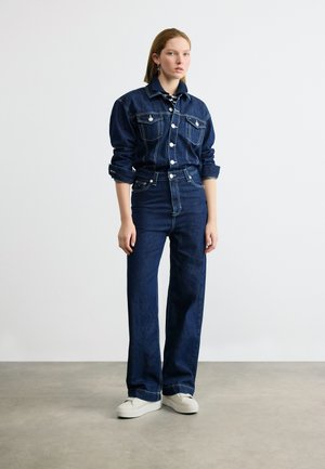 Junge Frau steht, trägt eine dunkelblaue Jeansjacke und hoch taillierte, weit geschnittene Jeans mit weißen Sneakern auf grauem Boden.