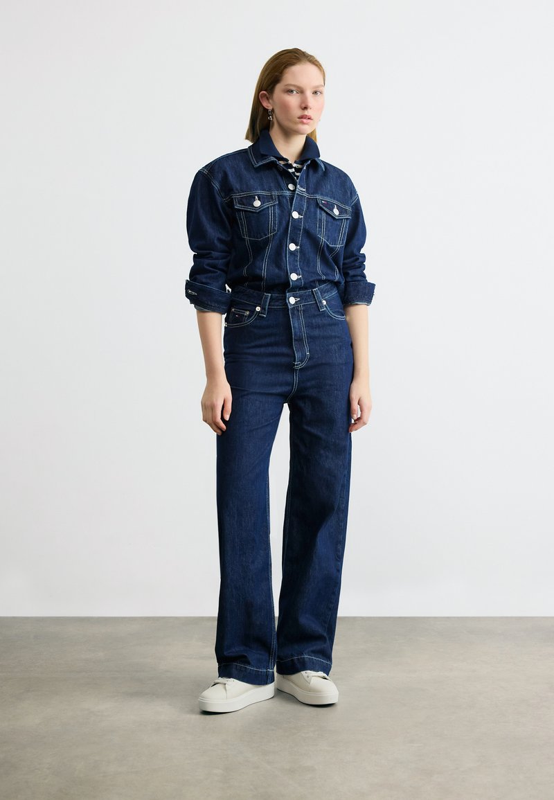 Jeune femme debout portant une veste en jean bleu foncé et un jean taille haute à jambes larges, avec des baskets blanches sur un sol gris.