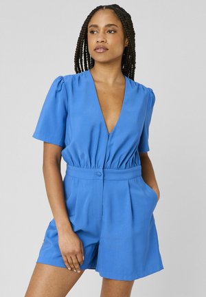 Femme aux cheveux nattés portant une combinaison courte à manches courtes bleu vif avec un décolleté en V profond et les mains dans les poches, debout devant un fond uni.