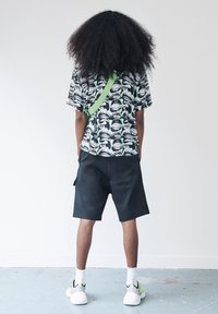 Person mit lockigem Haar, der mit dem Rücken zur Kamera steht, trägt ein schwarz-weißes Shirt mit Augenmuster, schwarze Shorts, weiße Socken und weiße Sneaker mit neonfarbenen Details.