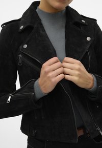 Černá suede motorkářská bunda s kovovými prvky a zipovými kapsami. Těsně padnoucí design zahrnuje špicatý límec a texturovanou látku.