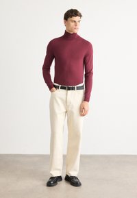 Mann iført en burgunder turtleneck, kremfargede bukser med et svart belte og svarte sko, står på et grått gulv mot en hvit vegg.