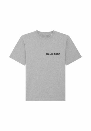 Jeune homme aux cheveux bruns bouclés porte un t-shirt gris clair avec le texte noir "YOU & ME TONIGHT" sur la poitrine gauche, assorti à un jean bleu clair.