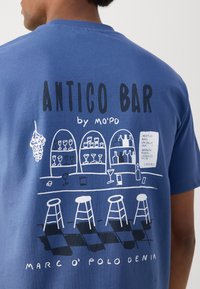Blauwe katoenen t-shirt met een afbeelding van een bar scène met glazen, drie krukken en tekst; zachte textuur en een losvallende pasvorm.