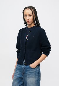 Καρδινάλιος πλεκτός σε ναυτικό μπλε με χρυσά κουμπιά, ραβδωτή υφή, στρογγυλή λαιμόκοψη και δύο μπροστινές τσέπες. Συνδυασμένος με ξεθωριασμένο μπλε τζιν.