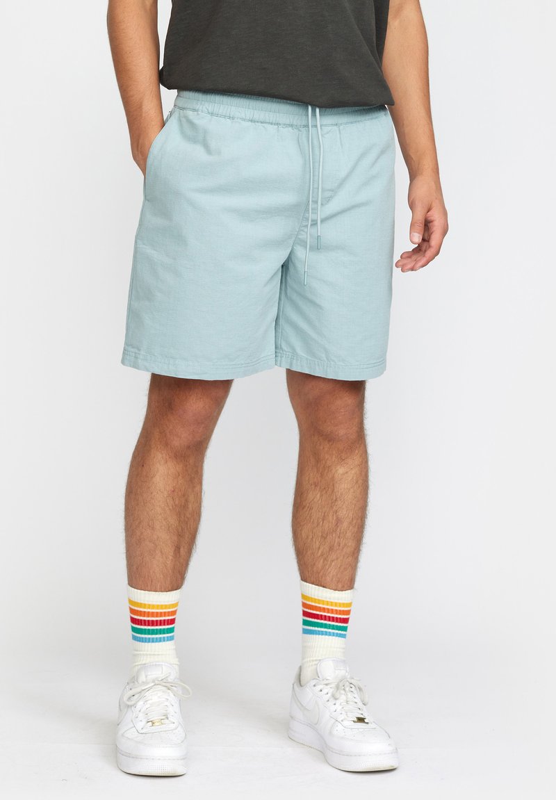 Lichtblauwe shorts met een elastische tailleband en drawstring. Gecombineerd met witte sneakers en gestreepte multi-kleurige sokken.