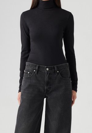 Femme portant un pull à col roulé noir ajusté et un jean en denim noir ample sur un fond uni.