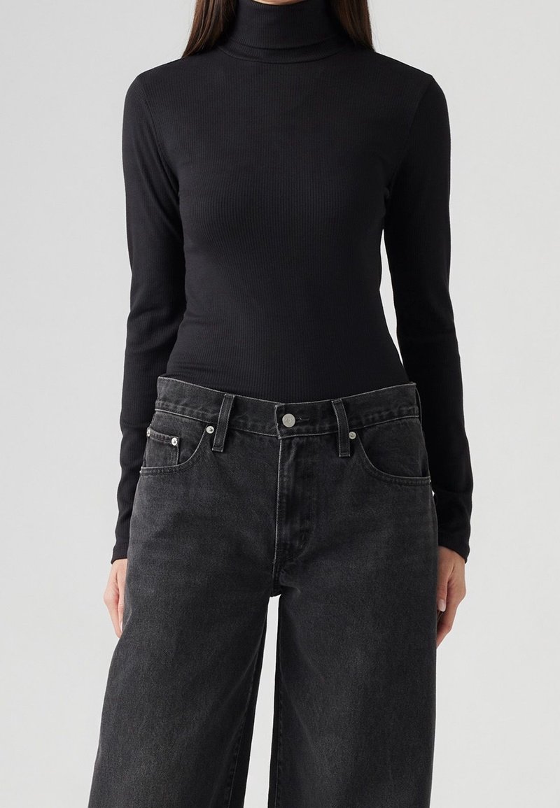 Femme portant un pull à col roulé noir ajusté et un jean en denim noir ample sur un fond uni.