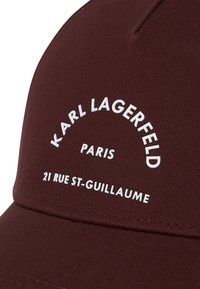 Tamsiai raudona beisbolo kepurė su baltais užrašais "KARL LAGERFELD PARIS 21 RUE ST-GUILLAUME" priekyje.