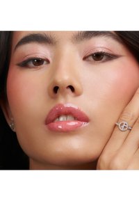 Anillo de plata con diseño circular y pequeños diamantes, llevado en una mano. Primer plano de un rostro con labios brillantes y maquillaje de color rosa claro.