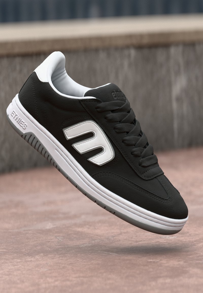 Sapatilha de couro preto com detalhes brancos, apresentando uma superfície texturizada, bico redondo e sola de borracha plana; logotipo na lateral.