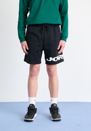 Person trägt schwarze Shorts mit Kordelzug und weißem Text, grünes Langarmshirt, weiße Socken und schwarze Turnschuhe und steht auf einem blauen Boden.