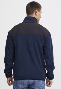 Marine fleecejack met zwarte schouderstukken, hoge kraag en textuurstof. Voorzien van lange mouwen en een relaxte pasvorm, geschikt voor casual gebruik.