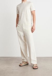 Man i en off-white t-shirt med kort ärm, lösa krämfärgade byxor och beige sandaler som står på ett ljusgrått golv mot en vit vägg.