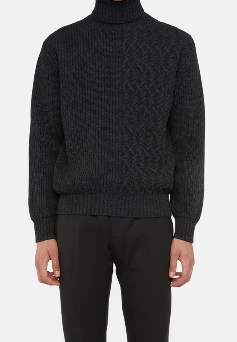 Homme portant un pull à col roulé gris foncé avec une texture côtelée et un panneau tricoté à motifs sur le côté droit, associé à un pantalon noir.