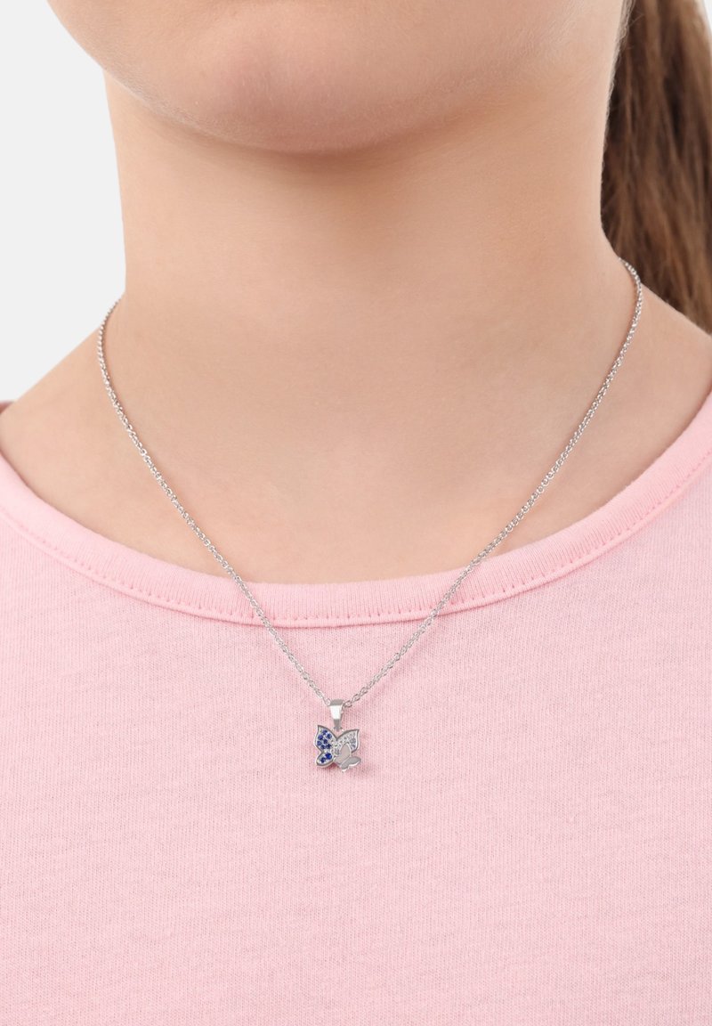 Collana in argento con pendente a forma di farfalla, decorata con gemme blu e trasparenti, indossata su una camicia rosa chiaro.