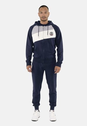 Homme debout portant un survêtement bleu marine avec un sweat à capuche à rayures diagonales grises et blanches et des baskets blanches sur fond blanc.