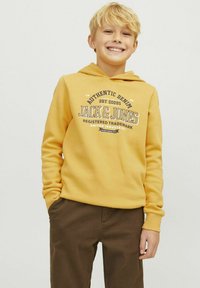 Felpa gialla realizzata in materiale morbido, con una grafica frontale che presenta il testo "JACK & JONES". Abbinata a pantaloni marroni, il design è casual e moderno.
