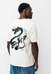 Fehér póló, amelynek hátulján egy nagy fekete sárkány motívum látható kék kiemelésekkel és piros japán felirattal. Normál fazon, rövid ujjú.