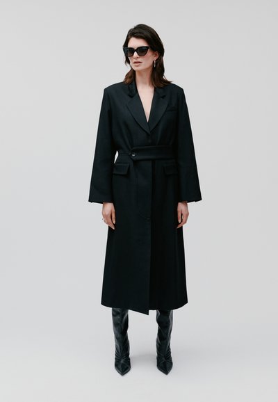 Cappotto lungo in stile blazer nero con vita cinturata, ampi revers e due tasche frontali. Indossato con stivali neri alti e appuntiti fino al ginocchio.