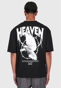 Fekete, oversized póló, amelyen egy fehér galamb grafika látható, körben börtöndróttal, alatta pedig a "HEAVEN" felirat és további szöveg.
