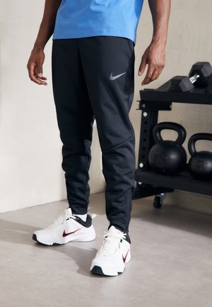 Person, der schwarze Nike-Jogginghosen und weiße Nike-Sneaker trägt, steht in der Nähe von Kettlebells und Kurzhanteln auf einem Fitnessstudio-Regal.