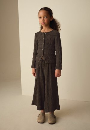 Mladé dievča stojí, oblečené v tmavohnedom textúrovanom krátkom svetri na gombíky a zodpovedajúcich širokých nohaviciach s béžovými slip-on topánkami, na neutrálom pozadí.